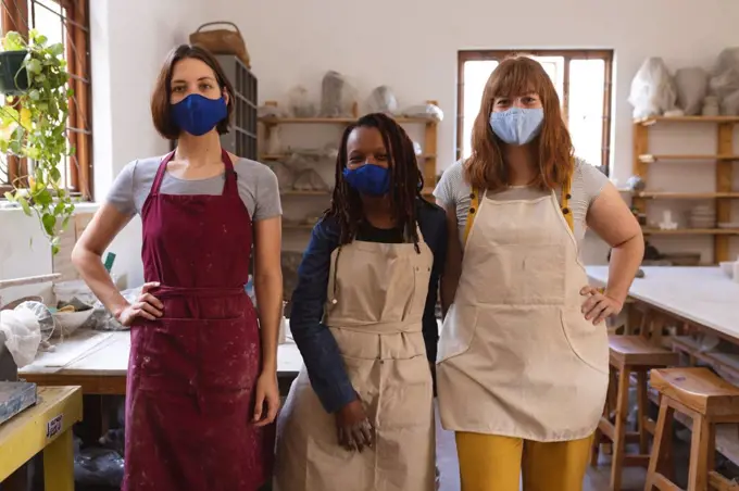 Two caucasian and one mixed race female potters in face masks working in pottery studio. wearing aprons, looking at a camera. small creative business during covid 19 coronavirus pandemic.