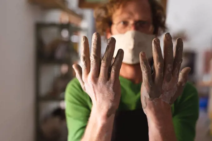 Caucasian male potter in face mask working in pottery studio. showing his dirty hands to the camera. small creative business during covid 19 coronavirus pandemic.