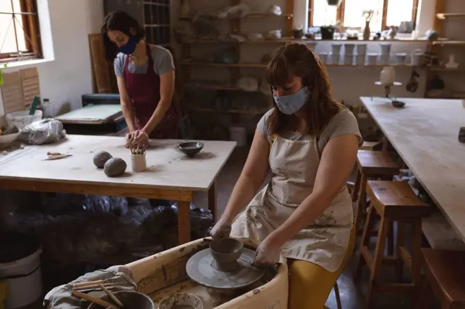 Two caucasian female potters in face masks working in pottery studio. wearing apron, working at a potters wheel and working table. small creative business during covid 19 coronavirus pandemic.