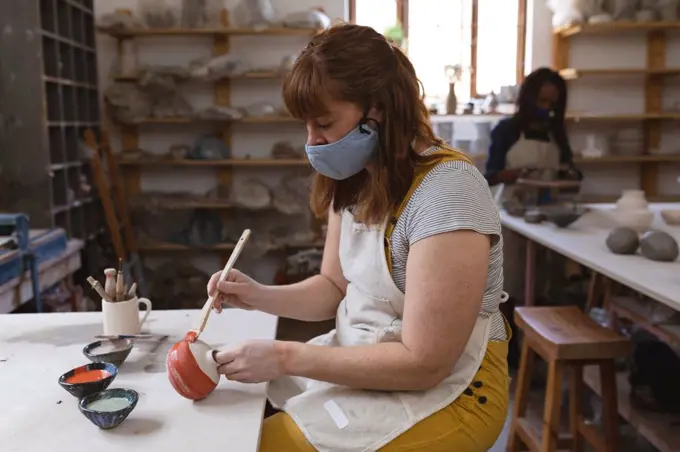 Caucasian female potter in face mask working in pottery studio. wearing apron, working at a working table, painting a plate. small creative business during covid 19 coronavirus pandemic.
