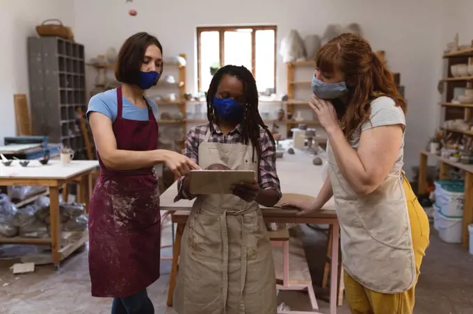 Two caucasian and one mixed race female potters in face mask working in pottery studio. wearing aprons, looking at digital tablet. small creative business during covid 19 coronavirus pandemic.