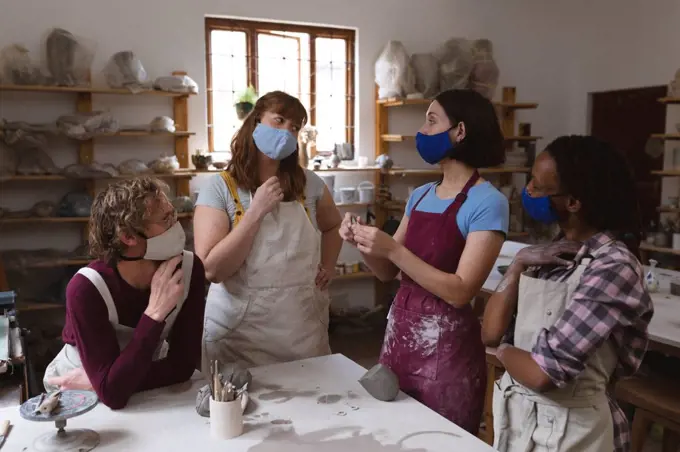 Multi-ethnic group of potters in face masks working in pottery studio. wearing aprons, talking. small creative business during covid 19 coronavirus pandemic.