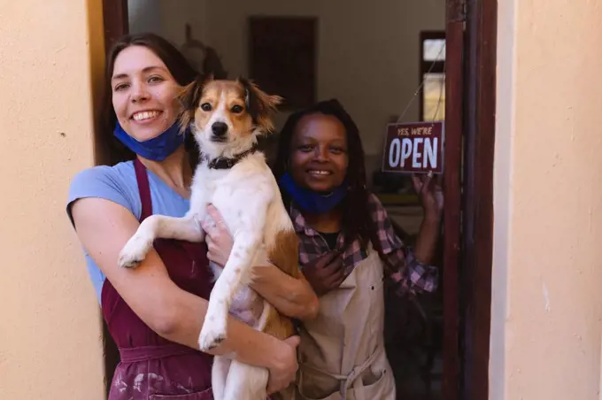 Portrait of caucasian and mixed race women at pottery studio, holding a puppy. small creative business during covid 19 coronavirus pandemic.