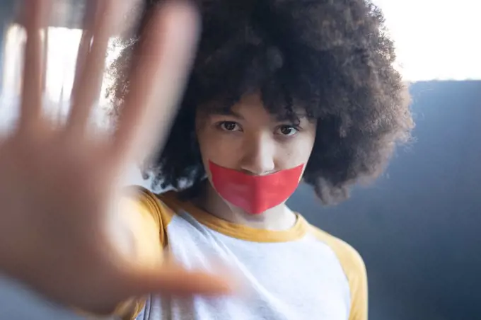 Mixed race woman with tape on mouth looking at camera holding her arm straight. gender fluid lgbt identity racial equality concept.