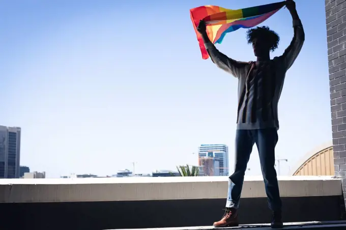 Mixed race man standing on rooftop holding rainbow flag. gender fluid lgbt identity racial equality concept.