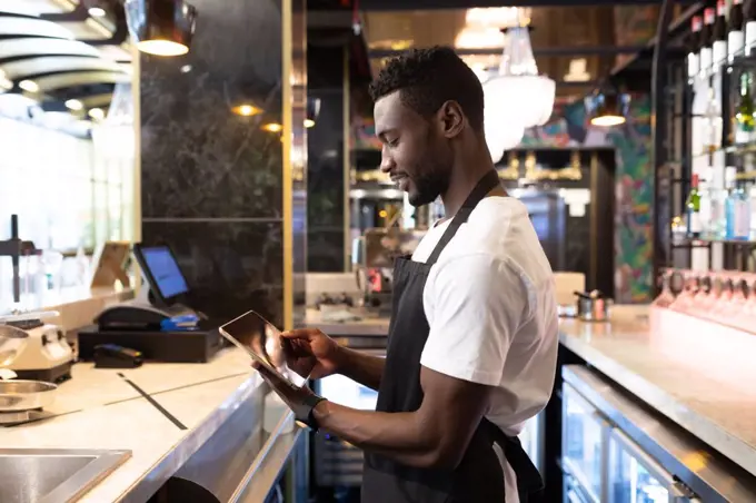 African american male barista standing behind a bar using a digital tablet and smiling. independent small business owner.