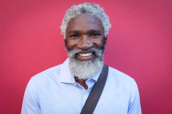 Portrait of african american senior man with beard standing against red wall in the street smiling. digital nomad out and about in the city.