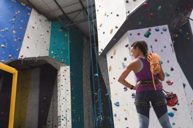 Caucasian woman with rope over her shoulder preparing for climb at indoor climbing wall. fitness and leisure time at gym.