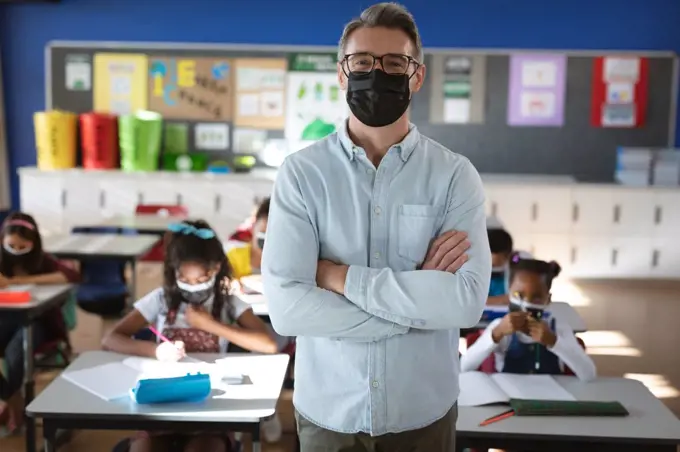 Portrait of caucasian male teacher wearing face mask standing in the class at elementary school. hygiene and social distancing at school during covid 19 pandemic