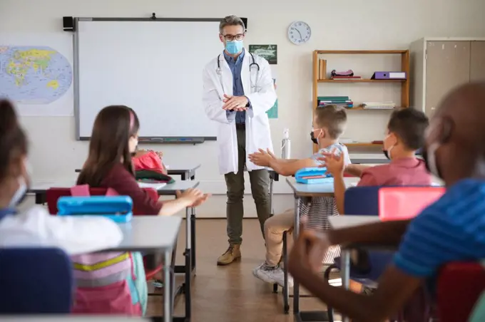Caucasian male doctor showing how to use hand sanitizer to group of diverse students at school. health protection and safety at school during covid-19 pandemic concept