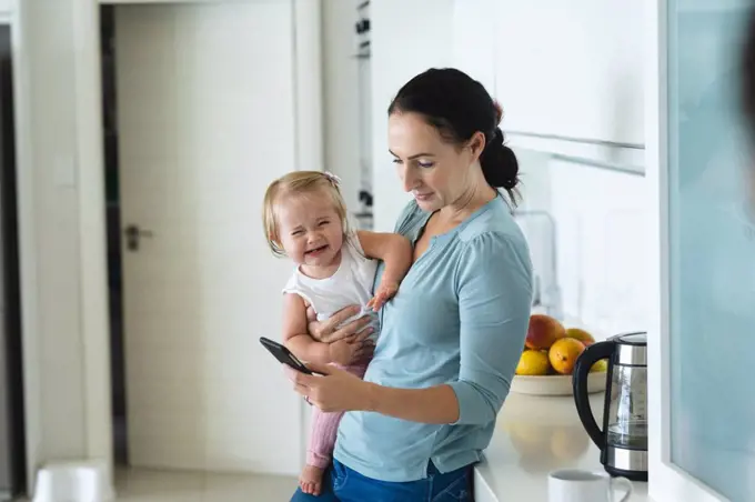 Caucasian mother holding her baby using smartphone in the kitchen at home. motherhood, love and baby care concept