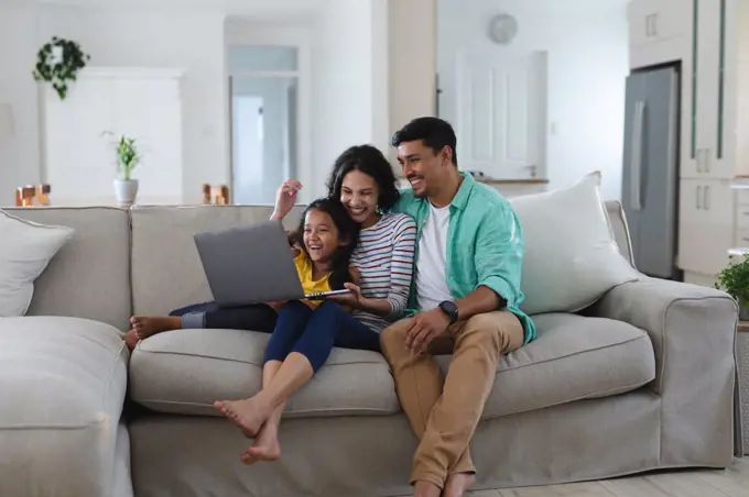 Smiling hispanic mother, father and daughter sitting on couch using laptop together. family spending time together at home.