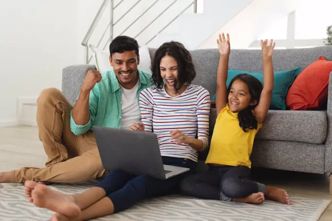 Cheering hispanic mother, father and daughter sitting on living room floor using laptop together. family spending time together at home.