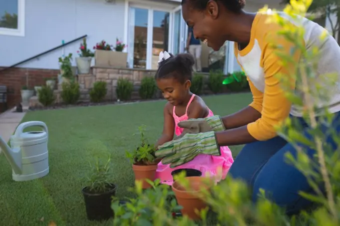 Happy african american mother and daughter kneeling tending to potted plants in garden. family spending time together at home.