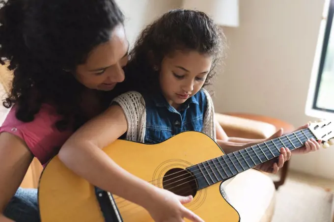 Mixed race mother and daughter sitting on sofa and playing guitar. domestic lifestyle and spending quality time at home.