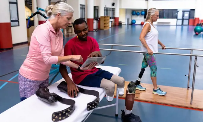 Side view of Caucasian active senior female trainer and disabled African-american man discussing over digital tablet in sports center. Sports Rehab Centre with physiotherapists and patients working together towards healing