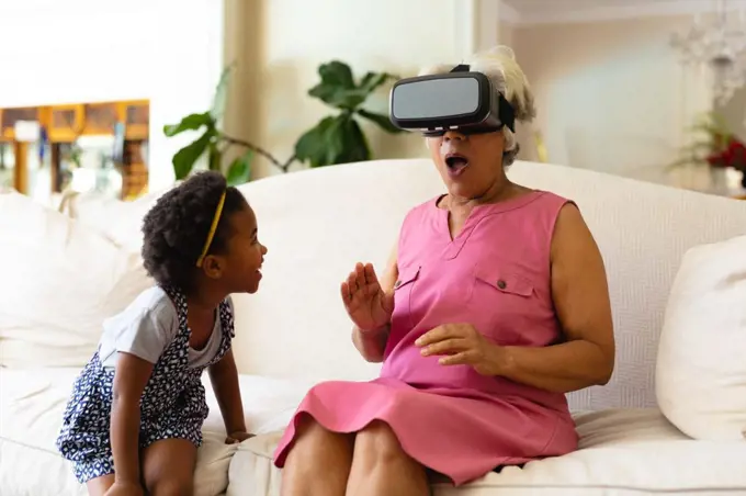African american girl looking at her grandmother wearing vr headset sitting on the couch at home. virtual reality and futuristic technology concept, unaltered.