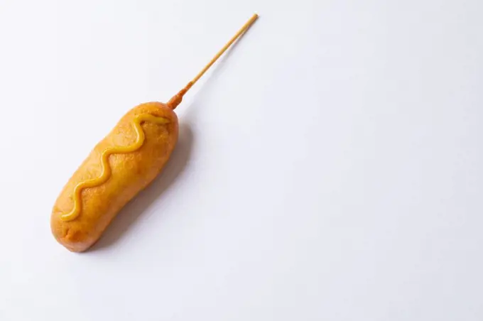 Close-up of corn dog with mustered sauce over skewer on white background with copy space. unaltered, copy space, unhealthy food, meat, sausage and fried food.