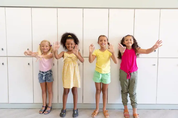 Portrait of cheerful multiracial elementary schoolgirls gesturing while standing against lockers. unaltered, childhood, together, education, enjoyment and back to school concept.