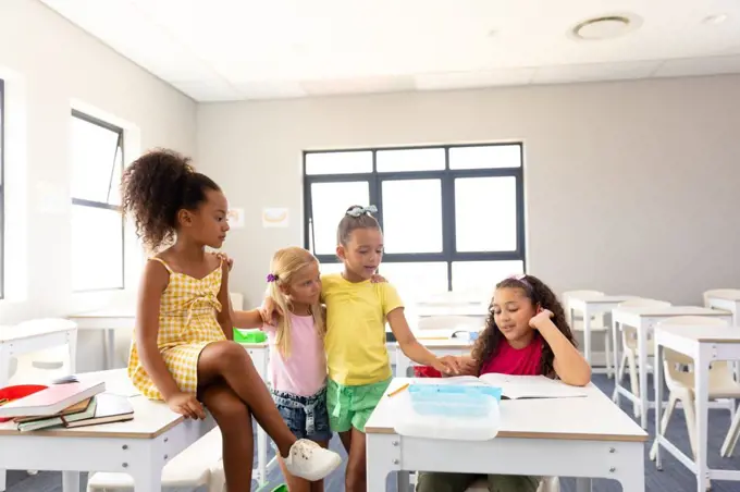 Multiracial elementary schoolgirls looking at female classmate reading book at desk in classroom. unaltered, childhood, learning, education and back to school concept.