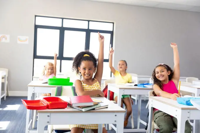 Smiling elementary schoolgirls raising hands while sitting at desk during class in school. unaltered, childhood, education, intelligence and back to school concept.