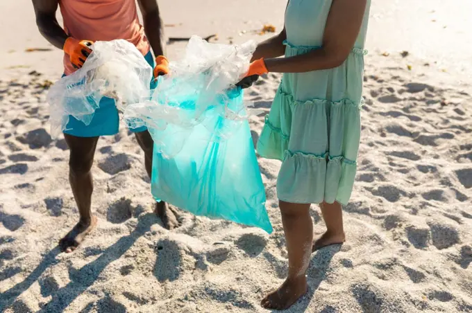 Low section of african american couple collecting plastic garbage in bag at beach on sunny day. unaltered, togetherness, responsibility and environmental issues concept.