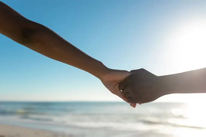 Cropped image of african american couple holding hands at beach against sky on sunny day. unaltered, lifestyle, love, togetherness and holiday concept.