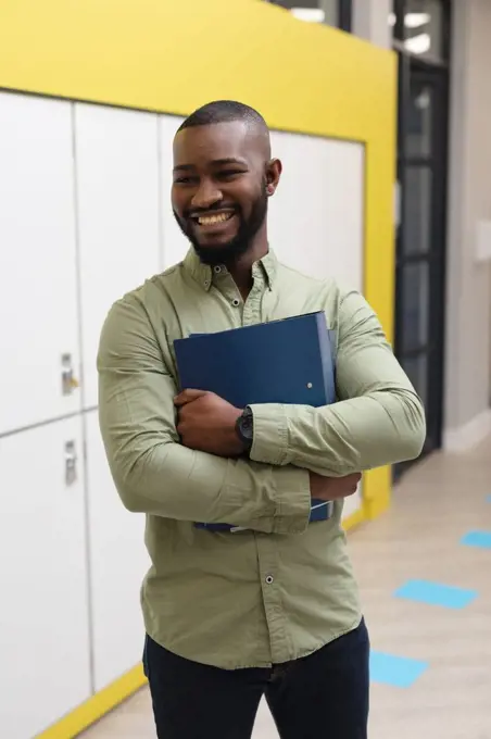 Smiling african american young male teacher with files looking away while standing in corridor. unaltered, education, occupation and school concept.