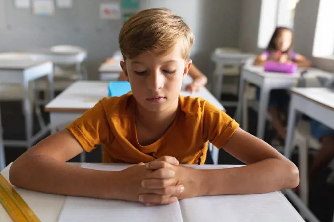 Caucasian elementary schoolboy with hands clasped studying at desk in classroom. unaltered, education, learning, childhood, studying and school concept.