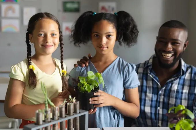 Portrait of biracial elementary schoolgirls with plant standing by african american young teacher. unaltered, education, childhood, teaching, science, stem and school concept.