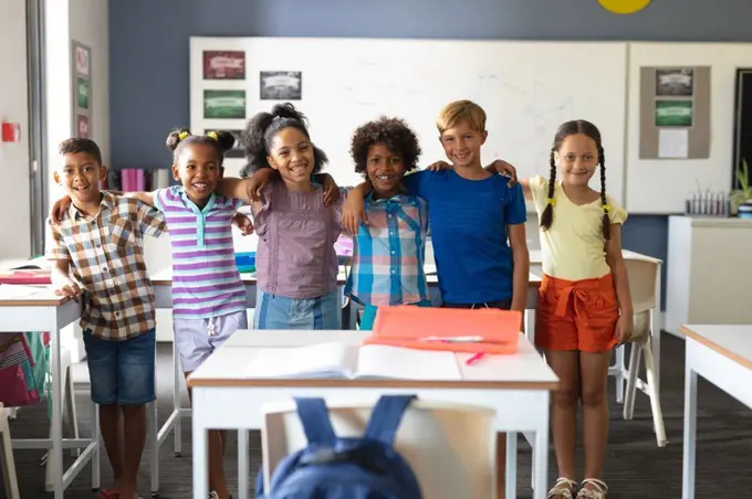 Portrait of smiling multiracial elementary school students standing with arm around in classroom. unaltered, education, childhood, learning, together and school concept.