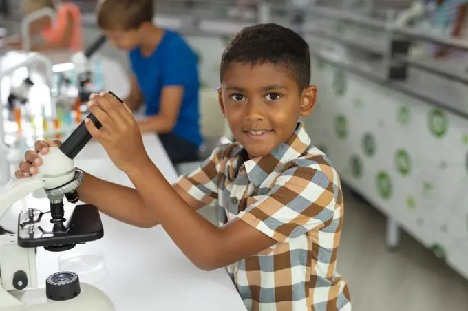 Portrait of biracial elementary schoolboy holding microscope while sitting at desk in laboratory. unaltered, education, childhood, learning, science, stem and school concept.