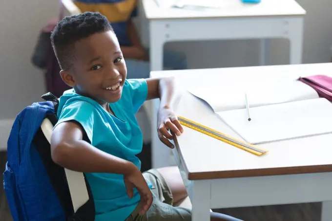 Portrait of smiling african american elementary schoolboy sitting on chair at desk in classroom. unaltered, education, childhood, learning, studying, book and school concept.