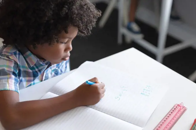 High angle view of african american elementary schoolboy writing on book at desk in classroom. unaltered, education, childhood, learning, studying and school concept.