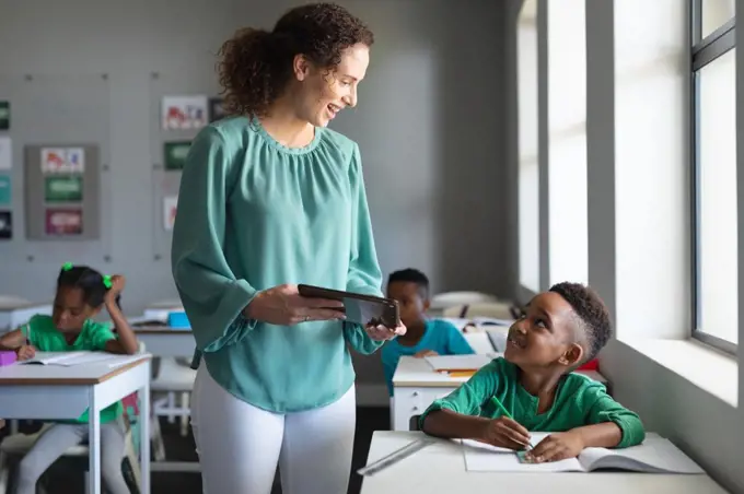 Smiling caucasian young female teacher showing digital tablet to african american elementary boy. unaltered, education, childhood, occupation, technology, teaching and school concept.