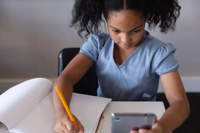 High angle view of biracial elementary schoolgirl using digital tablet while writing on book at desk. unaltered, education, learning, studying, concentration, wireless technology and school concept.