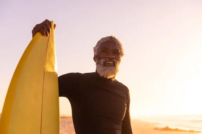 Smiling african american bearded senior man with surfboard standing at beach against clear sky. sunset, copy space, unaltered, retirement, aquatic sport, holiday and active lifestyle concept.