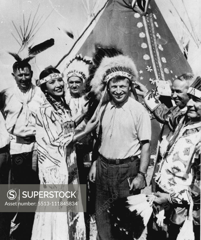 Stock Photo: 5513-111845834 Red Gets Redskin War Bonnet -- Miss Mary Louise Defender, Miss Indian II of Fort Yates, N.D., adjusts a war bonnet on the head of Yuri Golubash, deputy minister of state farms for the Soviet Union. Golubash and two other touring Russians looked in on the All-American Indian days celebration here while they were touring a number of Wyoming farms and ranches. The Russians said they had never seen Indians before, but this time they got a good look, more than 3,000 Indians are attending the meeting.