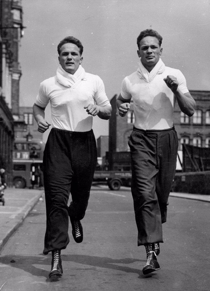 They'll Knock 'Em From The Old Kent Road -- Henry Cooper (on left) and his twin brother near their Old Kent doing a little roadwork near their Old Kent Road training quarters. Both are heavyweights. Jim Wicks, Manager of 20 year old boxer Henry Cooper, has refused an offer of $5,000 from film stars Donald Houston and Stanley Baker, for a half-share of the contract of Cooper, who comes from Bellingham, Kent. Henry is the identical twin brother of George Cooper, who is also a boxer. January 10, 1955. (Photo by Reuterphoto)