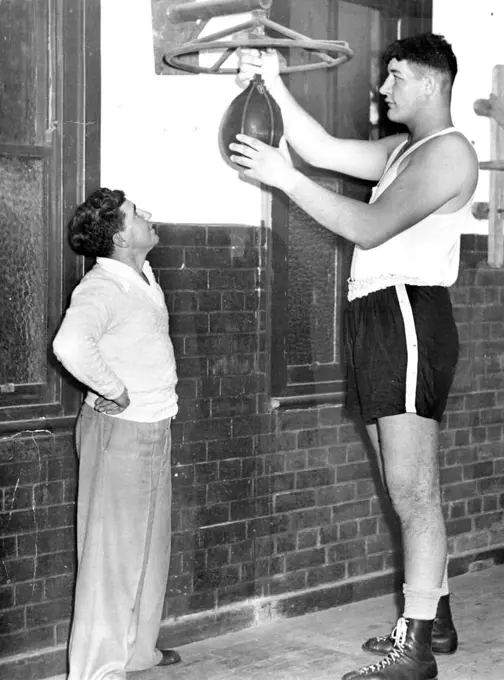 Ewart Potgieter, "The Vryheid Giant", screws a punch-ball into a special platform before starting training for his opening fight with Fred McCoy in Durban. Watching with a big grin is trainer Johnny Holt, a former S.A. bantam champion, who stands only 5ft 3in. September 16, 1955.