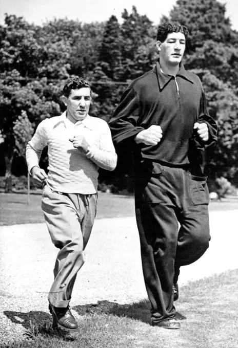 South African boxer Ewart Potgieter towers over his trainer John Holt in an exercise run at Brighton, England. Potgieter is 7ft 2in high and weighs 22½st. August 27, 1955.