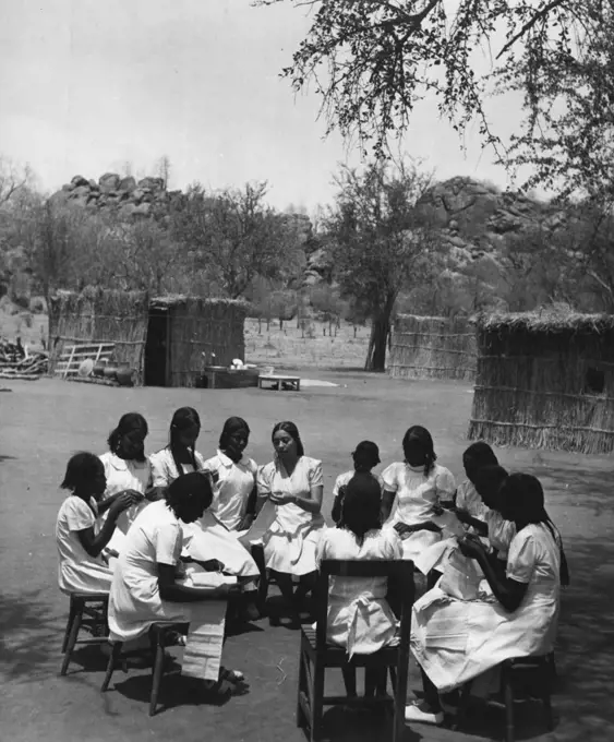 Sudanese Girls Train To Be Teachers.
They Learn In Grass Hut Classrooms Until New College Is Ready.
The sewing class sits in a circle round the teacher outside the grass huts which have been put up as temporary classrooms. These huts or tukls, are erected by Sudanese in a few hours. May 14, 1953. (Photo by Central Office Of Information).