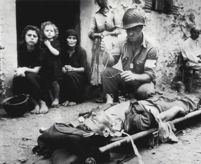 Holding Death At Bey -- Pvt. Harvey White, Minneapolis, Army hospital corpsman, holds death of Bey as he tensely administers blood plasma to a wounded Yank at first aid station in the street at San Agata, Sicily, which then was a half mile behind the front lines. What war means to the women in a war zone mirrored in the faces and hands of the women watching White fight with death. September 1, 1943. (Photo by AP Wirephoto).