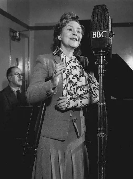 Annette Mills returns to broadcast a programme of her own songs in the BBC Forces Programme, on November 3rd, 1943. She is accompanied at the piano by Rex Burrows (in the background).
Annette Mills, the dancer who introduced the Charleston to Great Britain, and the composer of 'Boomps-a-daisy', has been in hospital for a year following a serious motor accident which occurred while she was driving between two R.A.F. stations to entertain the airmen. She suffered eight fractures, and is still dependent upon crutches and a sling. On November 4th, the day after this broadcast, she returns to hospital for what she hopes will be her final operation. November 03, 1943. (Photo by BBC).