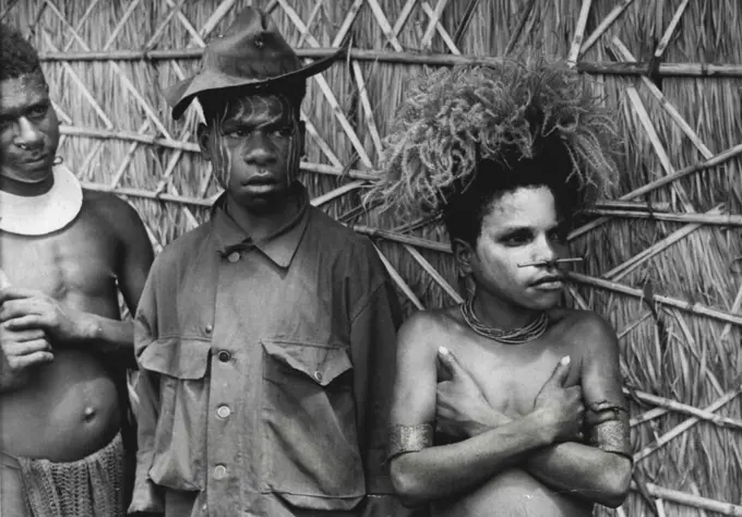 WI Favors western style clothing; his brother Kalano is conservative, sticks to a more traditional cut. But they both painted their faces from the same paint pot. They worked on the building of the kunai grass hut behind them. December 19, 1948.