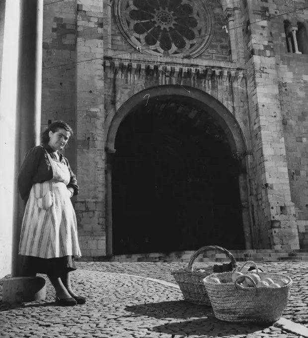 Cities of The World - Lisbon -- Part of Lisbon's scenery mush always include the lady vendors with their baskets of Madeira oranges, socks and other ware. This woman has set up shop in front of the city's 12th Century Cathedral. April 22, 1955. (Photo by United Press).
