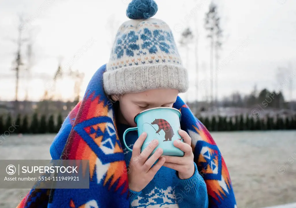 young boy wrapped in a blanket drinking hot chocolate outside