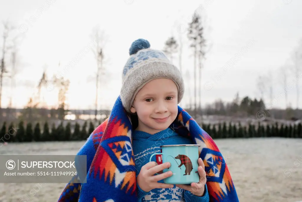 young boy smiling whilst wrapped in a blanket holding a hot chocolate