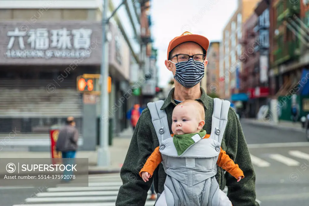 Father wearing face mask carrying daughter in baby carrier while crossing street in city during pandemic