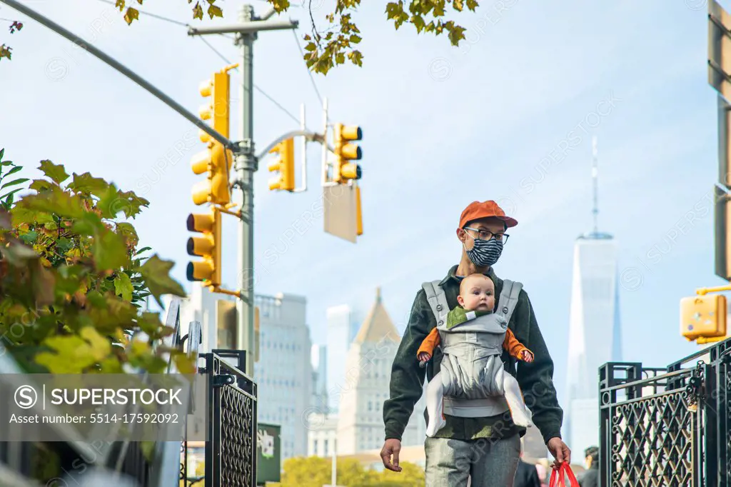 Father carrying cute daughter in baby carrier while walking in city during COVID-19